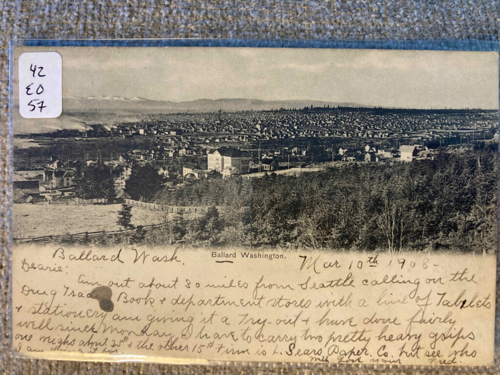 Scene of Ballard Washington--city and hills with mountains in distance. Text of handwritten message: “Ballard Wash. Mar 10th 1908 Dearie: Am out about 30 miles from Seattle calling on the Drug Trade Book + department stores with a line of Tablets + stationery am giving it a try-out + have done fairly well since Monday I have to carry two pretty heavy grips one weighs about 25# + the other 15# Firm is L. Sears Paper Co. but see who I am doing to for With love your Fred”