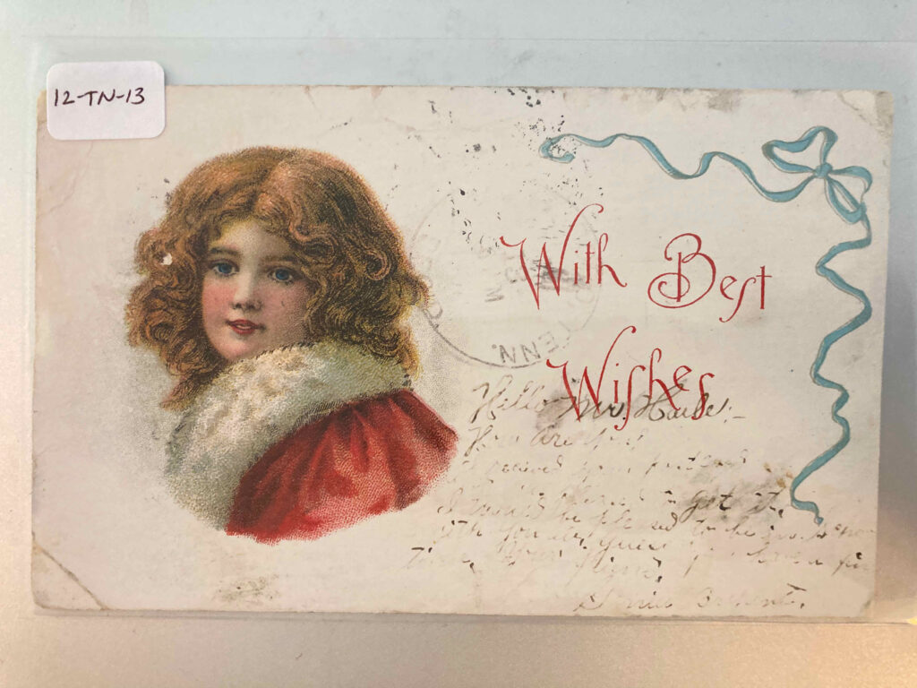 Head and shoulders of a woman in a fur-lined coat. Ribbon. Printed message "With Best Wishes." Inscription mostly illegible.